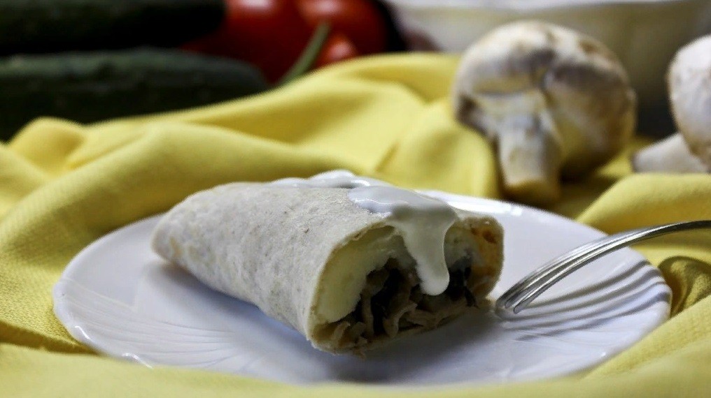 разрезанный блин с картофельным пюре и грибами, свернутый конвертом и политый сметаной, на белой тарелке с вилкой