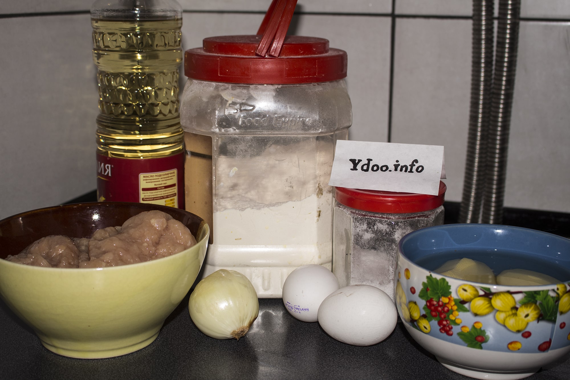 емкости с мукой и солью, два куриных яйца, очищенная луковица, несколько очищенных картофелин в пиале с водой, пиала с фаршем, бутылка растительного масла на кухонном столе